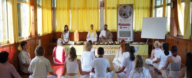 Group meditation or yoga class in a wooden hall with participants seated on mats, facing instructors at the front of the room.
