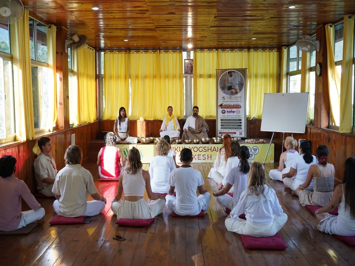 Group meditation or yoga class in a wooden hall with participants seated on mats, facing instructors at the front of the room.